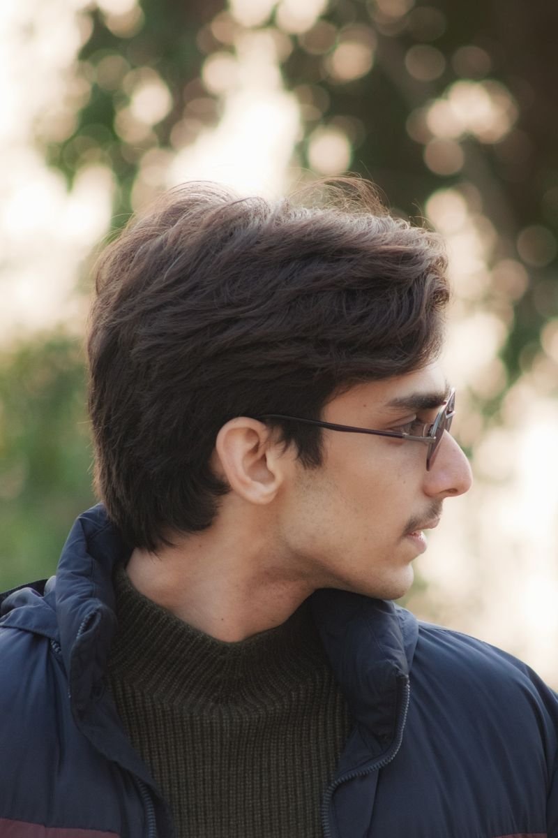 A thoughtful portrait of a young man wearing glasses and a winter coat, set against a blurred outdoor backdrop.